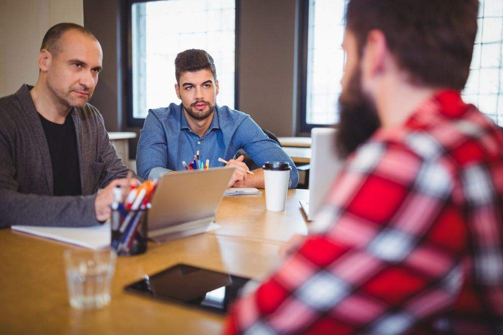 três profissionais trocam informações em uma reunião para ilustrar a comunicação assertiva nas empresas