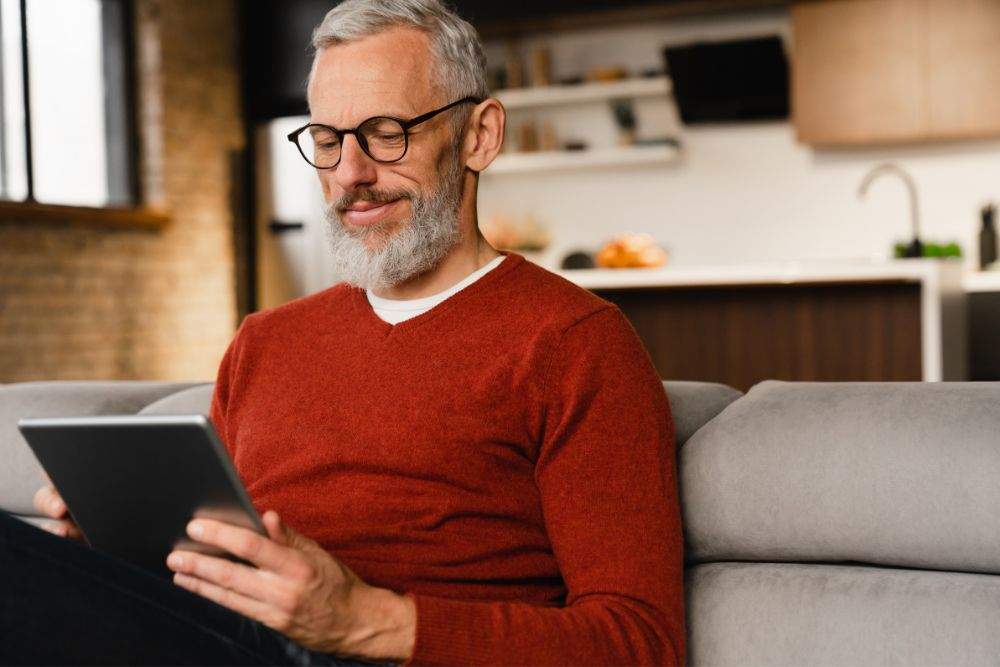 homem está lendo uma das newslettes para gestores em um tablet sentado ao sofá e expressãndo satisfação