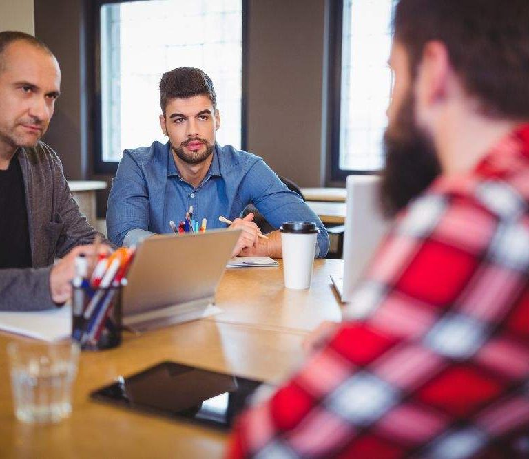 três profissionais trocam informações em uma reunião para ilustrar a comunicação assertiva nas empresas