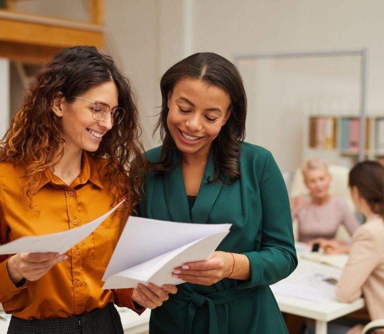 duas mulheres observam relatórios e estão contentes com os resultados da gestão por competências